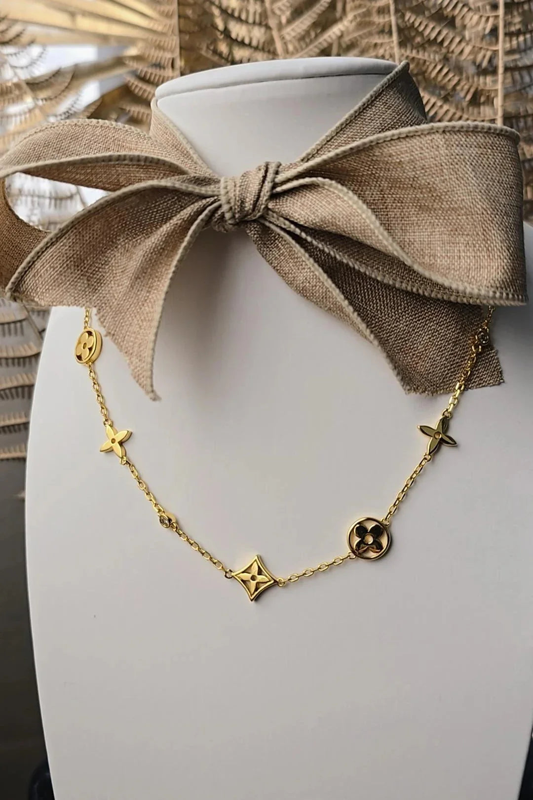 Gold charm necklace displayed on a white stand with a beige bow, Bliss Bymi jewelry