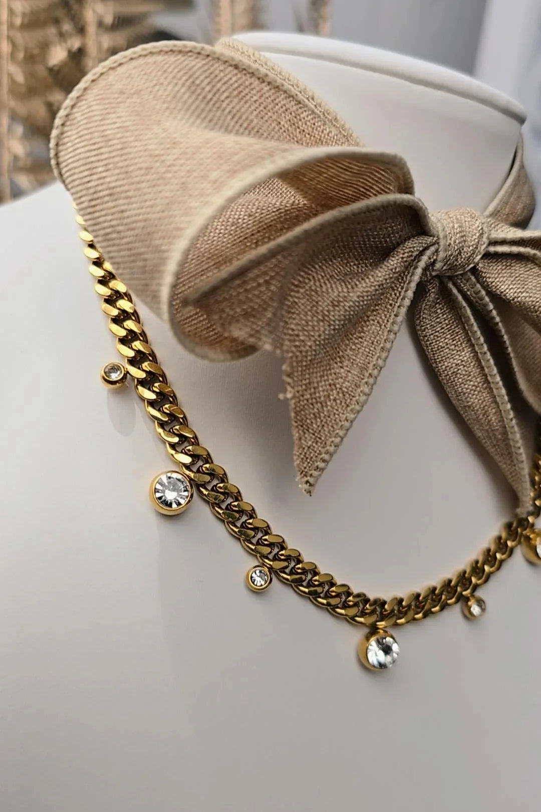 Gold chain necklace with crystal pendants displayed on a stand with a beige fabric bow, Bliss Bymi jewelry