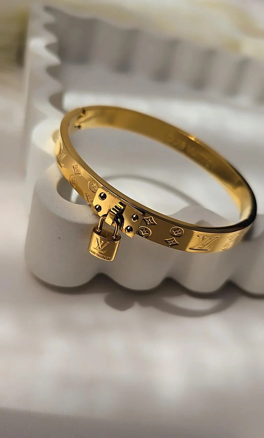 Gold bracelet with engraved details and small lock charm, displayed on a white jewelry holder.