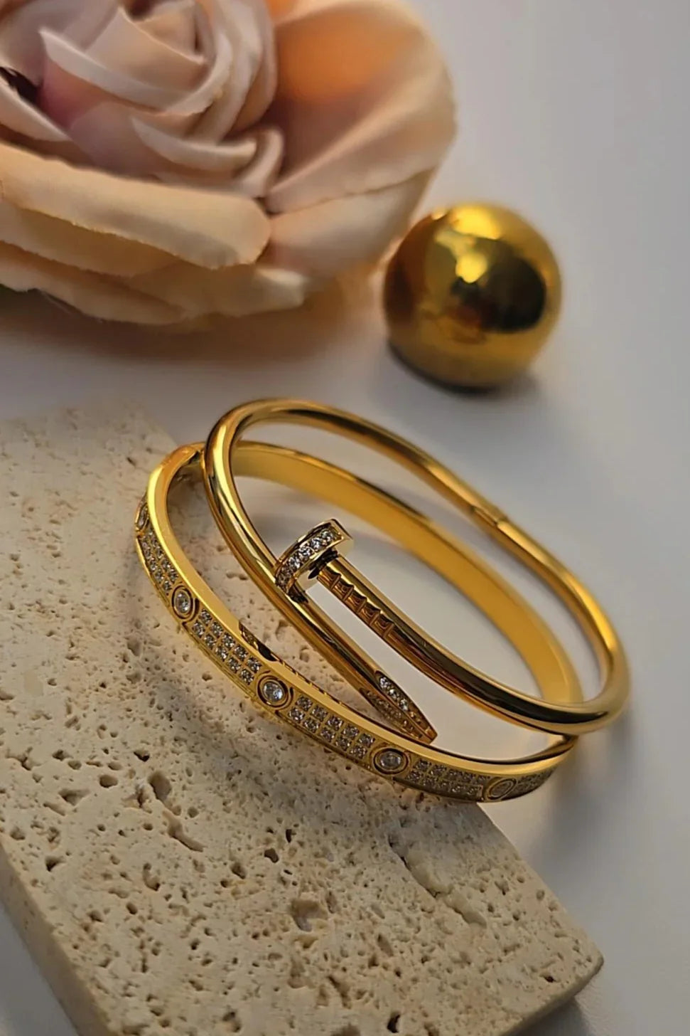 Gold bangle bracelets with crystal accents from Bliss Bymi, styled with a rose and stone background.