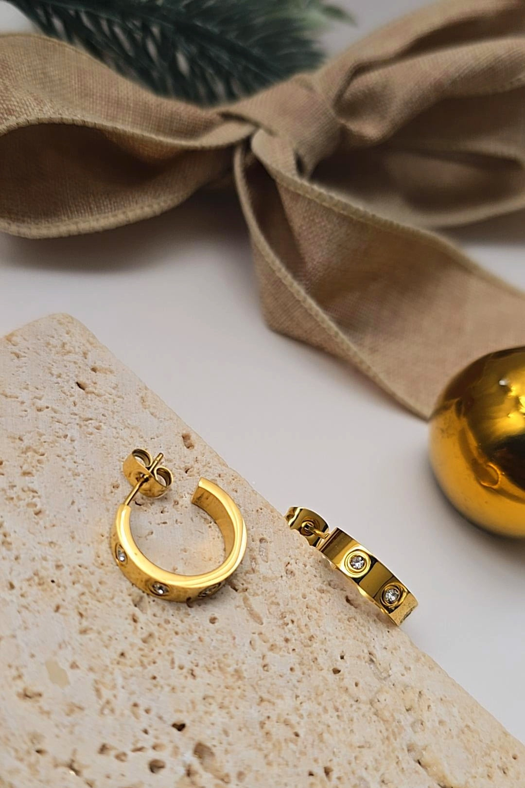 Gold hoop earrings on a textured surface with a beige ribbon and gold bell in the background.