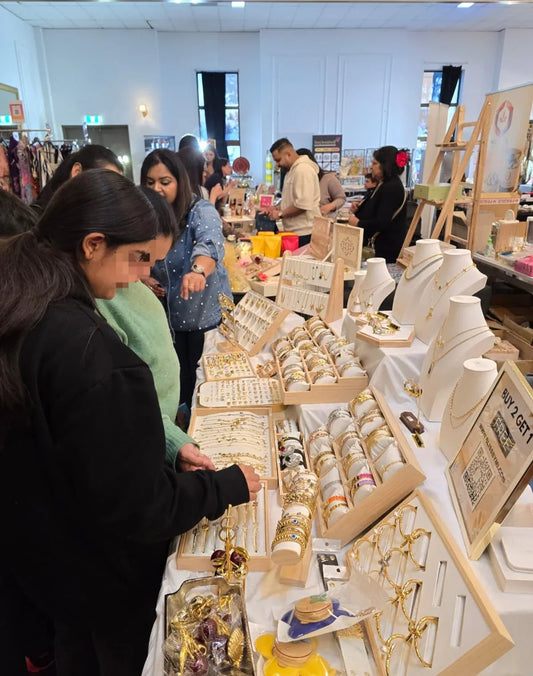 Shoppers browsing Blissbymi jewelry display with earrings, bracelets, and necklaces at a market stall.
